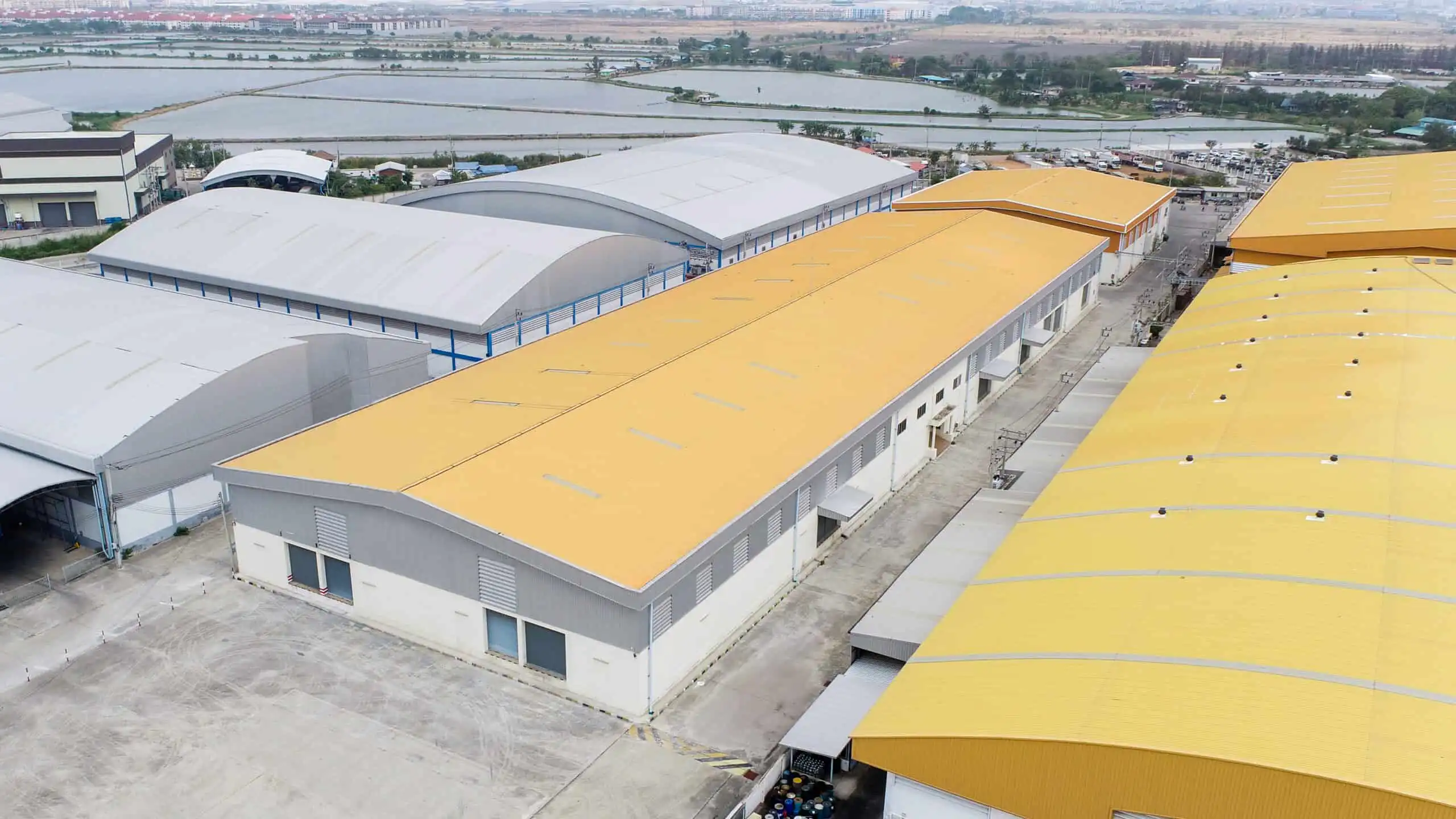 Warehouse and factory buildings with yellow and gray roofs, surrounded by paved areas and distant greenery.